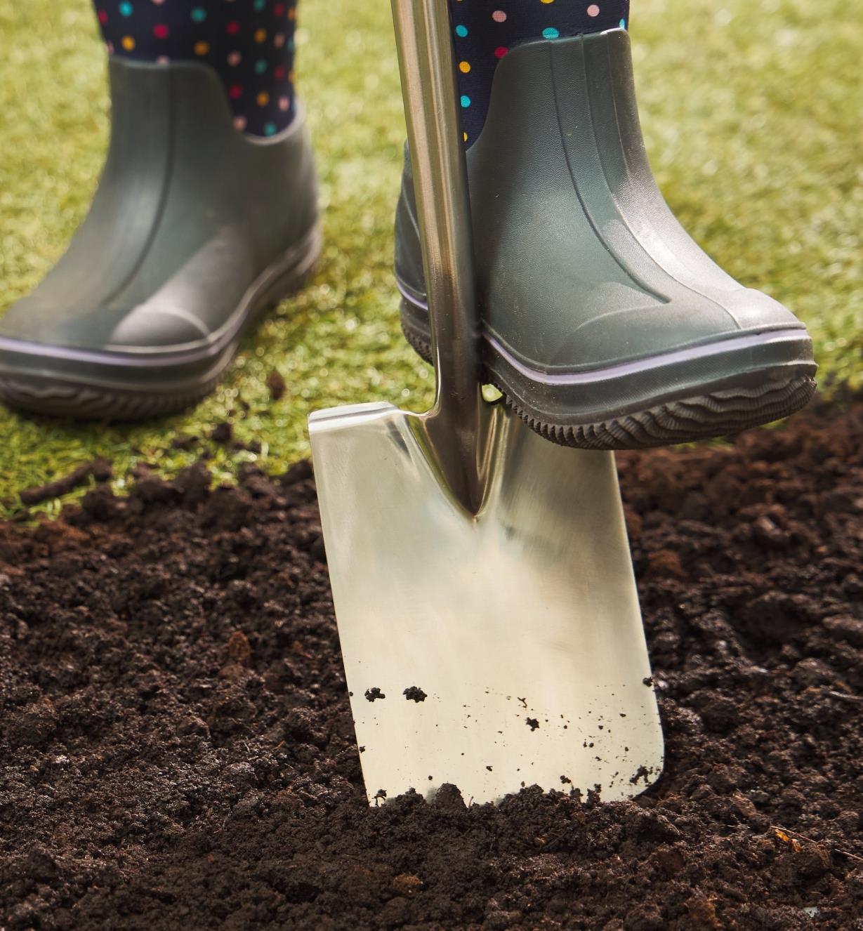 A foot wearing a rubber boot pushes the children’s digging spade into soil 