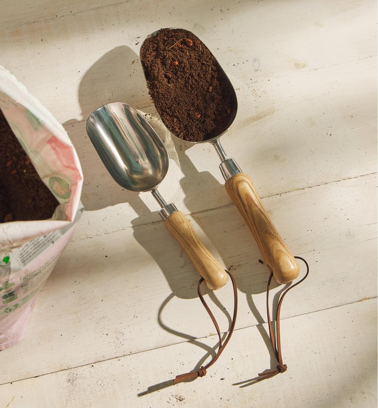 A 1 cup stainless-steel scoop filled with soil sits next to an empty ½ cup scoop on a wood surface