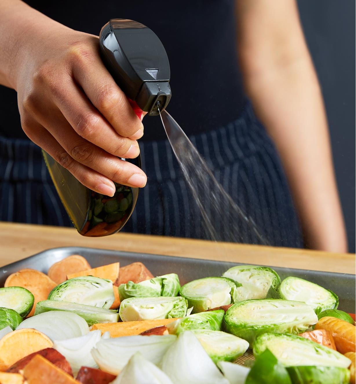 Using the oil sprayer to spray oil on sliced vegetables laid out on a baking pan