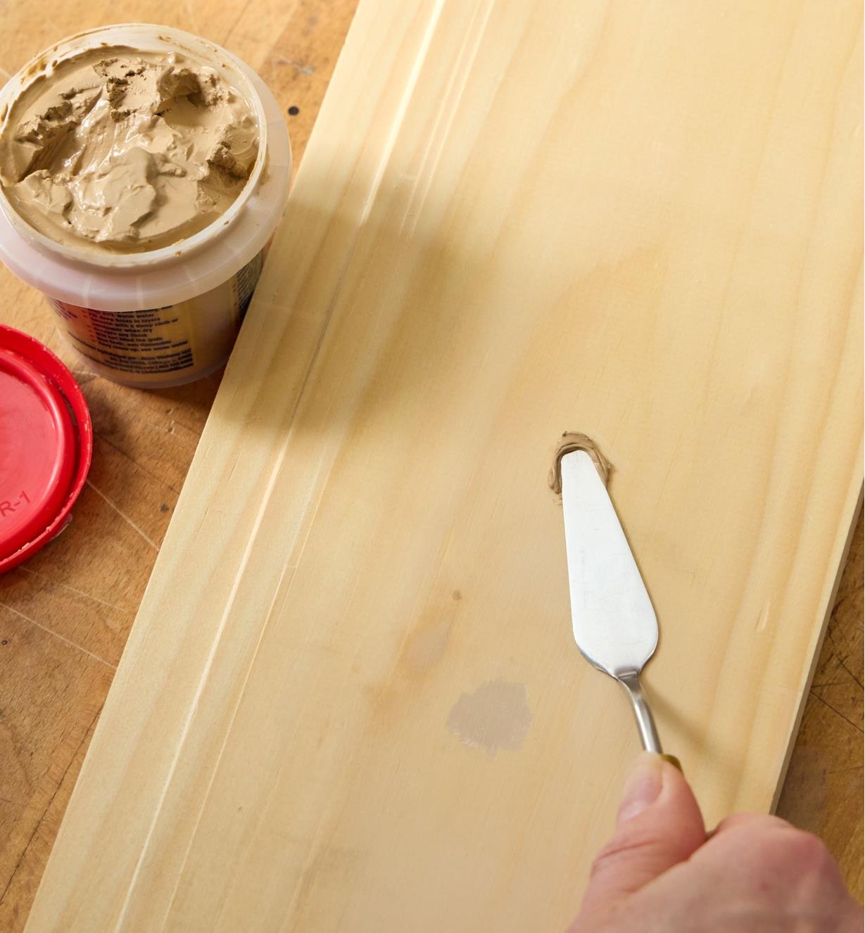 Personne appliquant du bouche-pores couleur bois avec une spatule sur une planche à côté d’un pot de bouche-pores ouvert