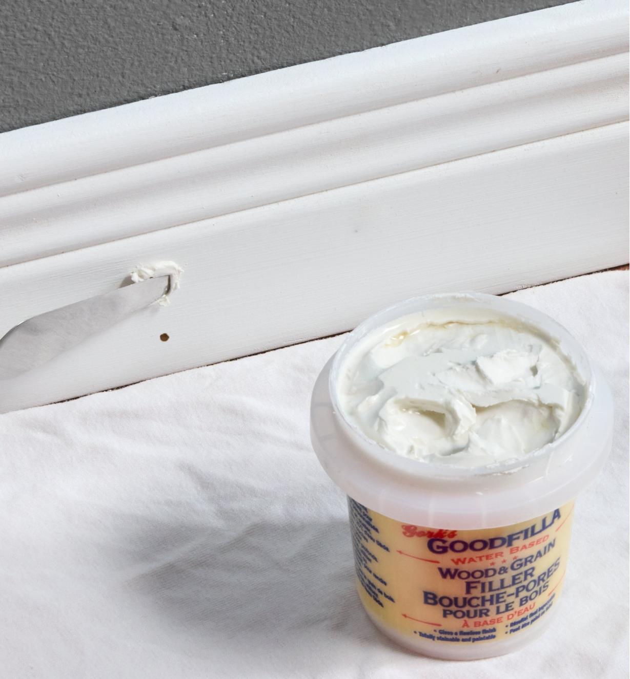 An open container of base/neutral wood filler next to a baseboard with filler applied to a pin hole