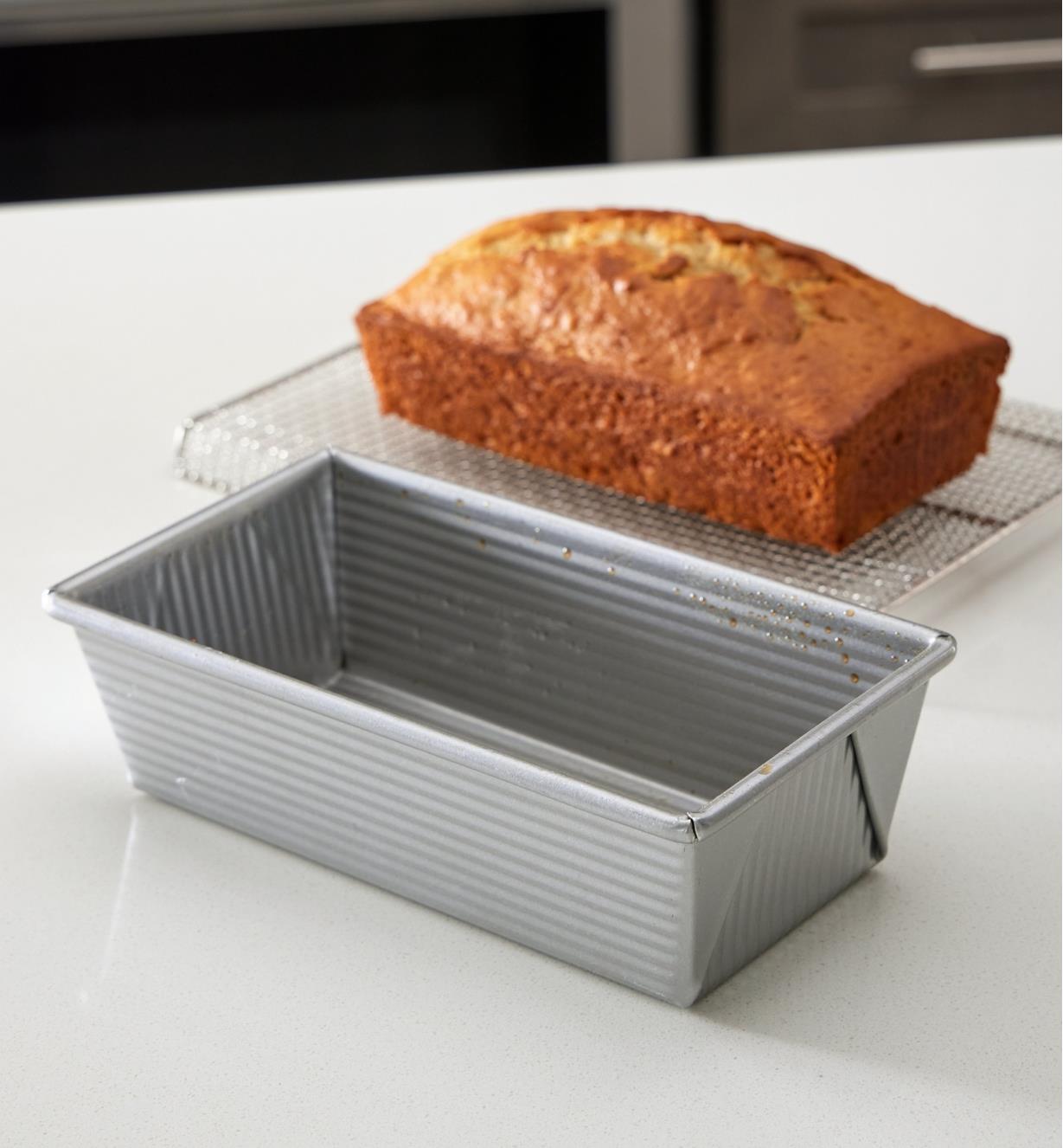 A loaf of bread sits on a cooling rack next to an empty load pan