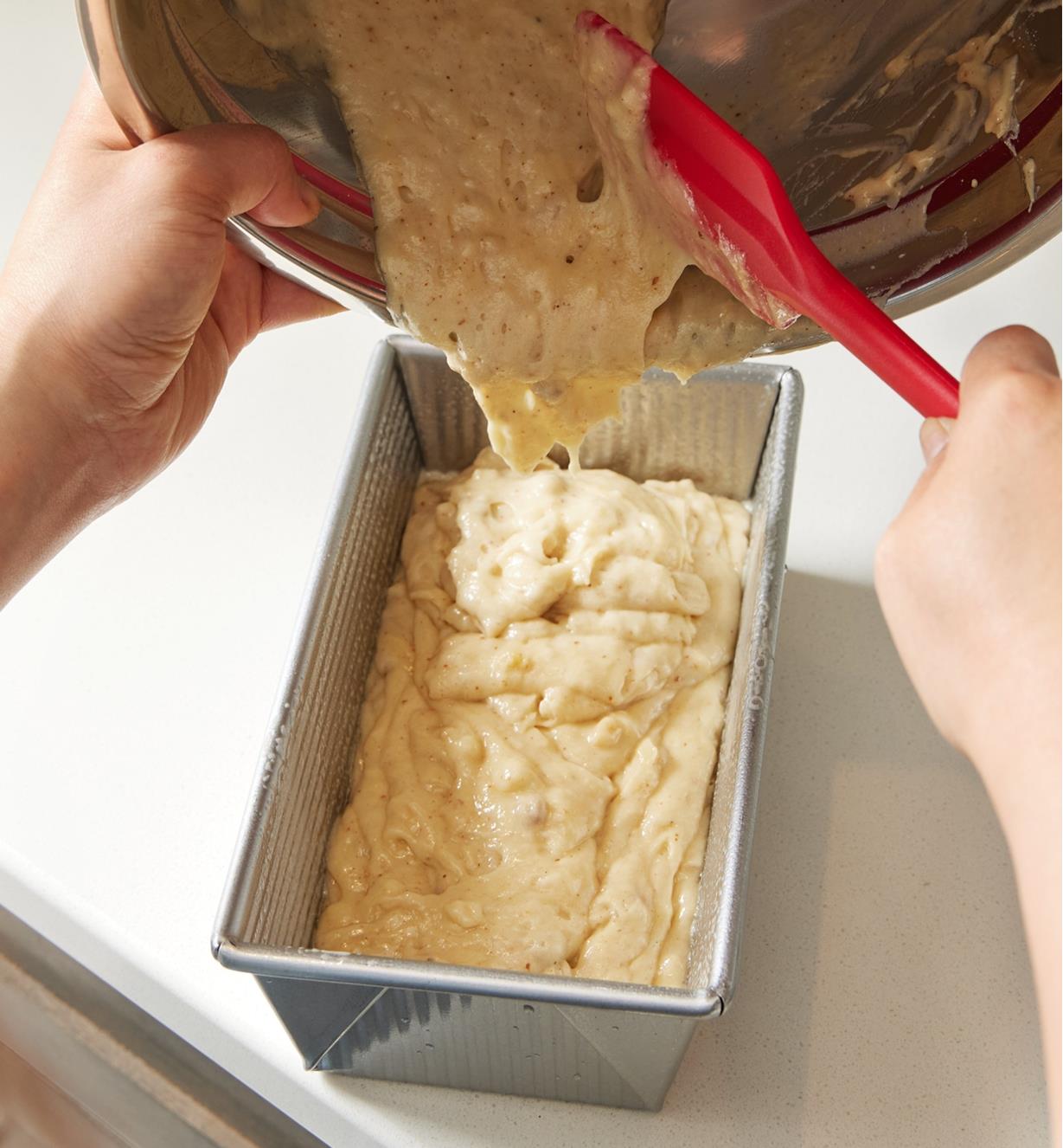 Bread batter being poured from a metal bowl into the loaf pan