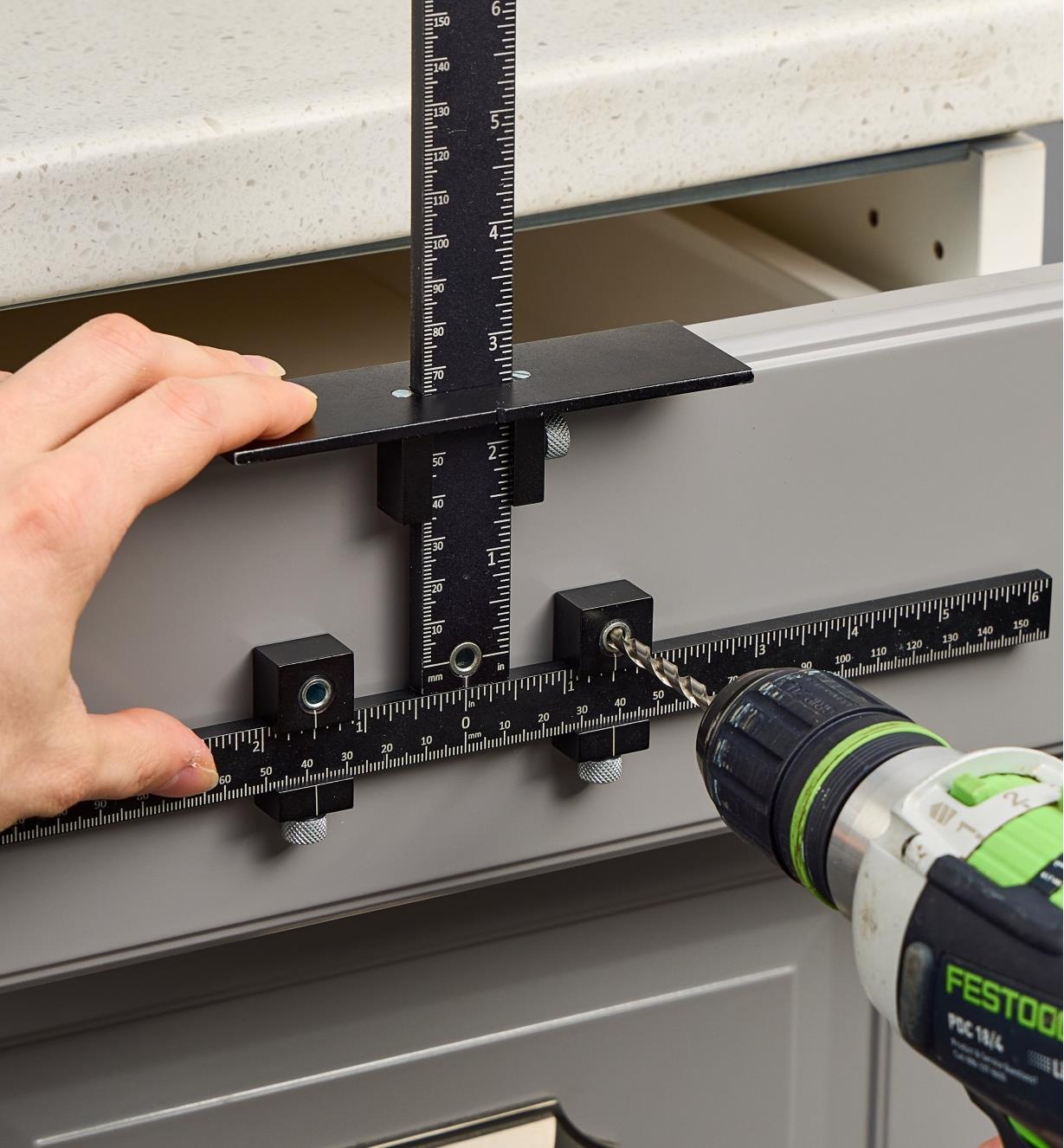 A cabinet hardware jig positioned on a drawer with a hole being drilled through the drill guide